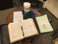 An open Bible and a book titled 'Becoming a Millionaire God's Way' are placed on a wooden table. There is a cup with the Starbucks logo, handwritten notes on a notecard, a pen resting on the Bible, and a black leather chair partially visible in the background.