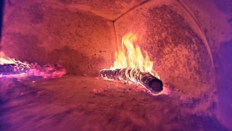 A brick oven interior displaying a large wooden log on fire, with flames visibly emanating from the log. The walls of the oven are curved and coated with soot, providing a rustic and warm ambiance.