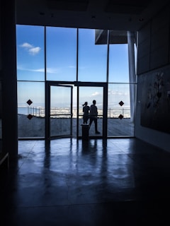 Large glass windows reveal an outside view of a wooden deck and the ocean beyond. Two silhouetted figures are standing near the window, with a clear blue sky and a few clouds visible. Inside, the floor reflects light, creating a contrasting dark interior.
