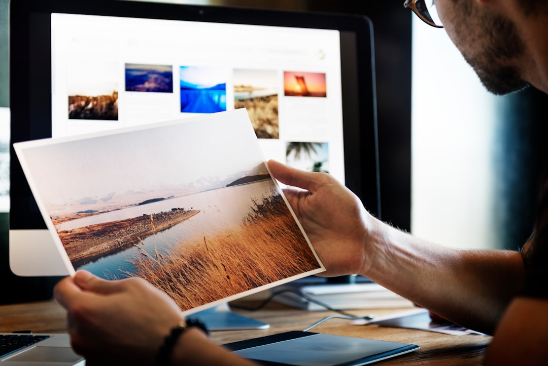 Man wearing glasses holding printed photo in front of computer work space