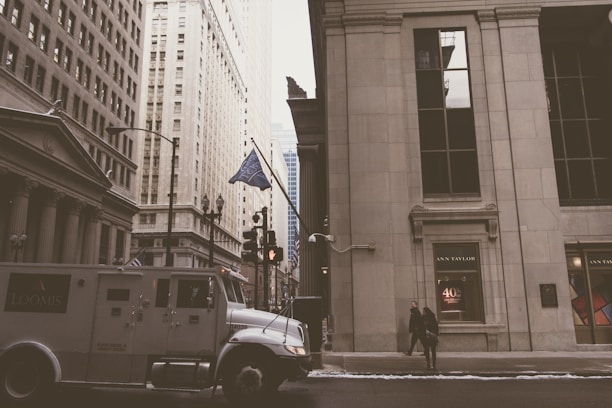 Tall buildings line a city street with a focus on a Loomis security truck in the foreground. Pedestrians are walking along the sidewalk, and flags are visible on the buildings. The architecture features classical columns and large windows, and there is an Ann Taylor storefront.