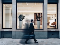 A man in a suit walks past a modern office building with large glass windows. Inside, there are two large potted plants, sleek wooden paneling, and a bright reception area with modern furnishings.