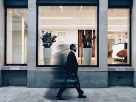 A man in a suit walks past a modern office building with large glass windows. Inside, there are two large potted plants, sleek wooden paneling, and a bright reception area with modern furnishings.