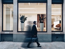 A man in a suit walks past a modern office building with large glass windows. Inside, there are two large potted plants, sleek wooden paneling, and a bright reception area with modern furnishings.