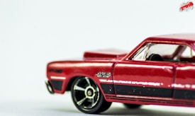 A close-up of a red toy car focusing on the side profile. The car features detailed elements such as black racing stripes, visible wheel rims, and a small decal with the text '426 HEMI'. The background is a soft white, accentuating the vivid color of the car.