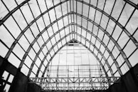 A large, ornate metal framework forms a series of arches, creating an open and spacious architectural structure. The grid-like pattern of the beams produces a striking geometric design against the cloudy sky.