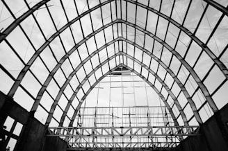 A large, ornate metal framework forms a series of arches, creating an open and spacious architectural structure. The grid-like pattern of the beams produces a striking geometric design against the cloudy sky.
