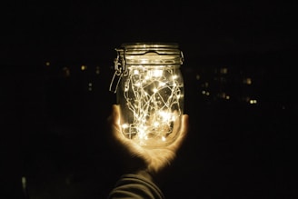 A hand holds a glass jar filled with glowing fairy lights, set against a dark background.