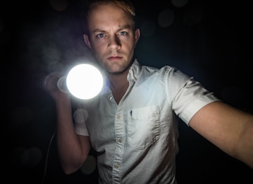 A person in a white collared shirt holds a bright flashlight directed towards the viewer, set against a dark background. Their expression is serious and focused.