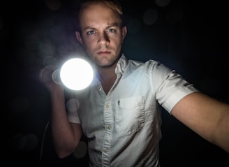 A person in a white collared shirt holds a bright flashlight directed towards the viewer, set against a dark background. Their expression is serious and focused.