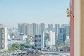 A cityscape with numerous high-rise buildings in the background and a person suspended on the side of a tall building in the foreground. The person appears to be painting or performing maintenance, wearing a safety harness and helmet.