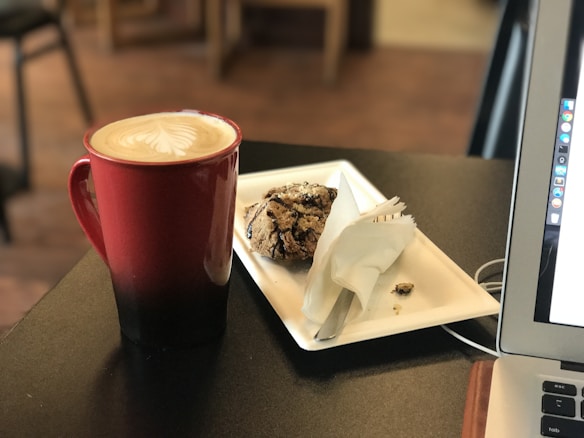 A red mug filled with coffee and topped with latte art is placed next to a white rectangular plate holding a baked pastry. A laptop is open to the right side of the table, with a partial view of its screen. The background includes a blurred view of furniture, suggesting the setting is a cafe.
