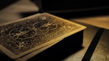 A deck of playing cards with intricate designs on the back, resting on a surface. The lighting is low and casts a warm, golden glow, highlighting the detailed patterns. The focus is on the textures and the beautiful craftsmanship of the card design.