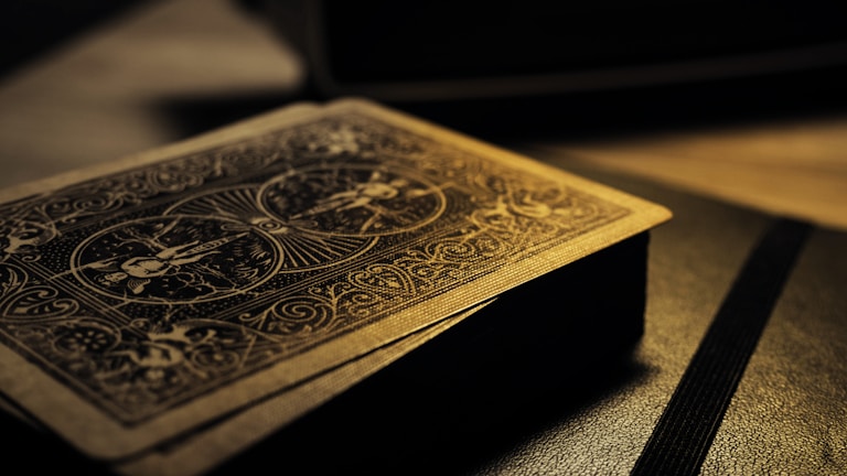 A deck of playing cards with intricate designs on the back, resting on a surface. The lighting is low and casts a warm, golden glow, highlighting the detailed patterns. The focus is on the textures and the beautiful craftsmanship of the card design.