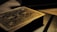 A deck of playing cards with intricate designs on the back, resting on a surface. The lighting is low and casts a warm, golden glow, highlighting the detailed patterns. The focus is on the textures and the beautiful craftsmanship of the card design.