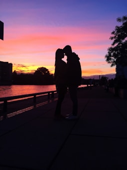 A silhouette of a couple stands close together, seemingly embracing or about to kiss, set against a vibrant sunset sky. The horizon displays rich hues of pink, purple, and orange, creating a romantic and serene atmosphere. Trees and buildings can be seen prominently in the foreground, along with a calm waterfront.