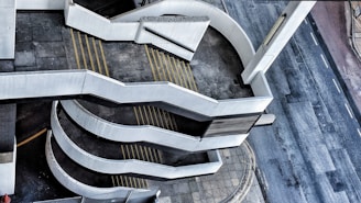 An empty multi-story car park ramp with a spiral design. The concrete structure is prominent, with yellow directional lines on the ground. Adjacent to the ramp is a quiet, empty street. The perspective offers an aerial view, emphasizing the geometric patterns formed by the ramps.