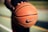 A detailed close-up of a hand holding a well-worn basketball with the Nike logo in focus. The blurred background suggests an outdoor basketball court with green and yellow hues, indicating lines and potential grassy areas.
