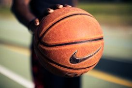 A detailed close-up of a hand holding a well-worn basketball with the Nike logo in focus. The blurred background suggests an outdoor basketball court with green and yellow hues, indicating lines and potential grassy areas.