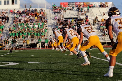 A group of American football players in yellow and white uniforms are lined up on a field, ready to kick off. The stands in the background are filled with spectators, some of whom are wearing green shirts.