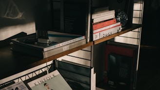 A wooden bookshelf filled with various books and magazines, some stacked and others neatly lined. An old-fashioned iPod and a pair of earbuds rest on top of a pile of books on the upper shelf. There is a clock and some decorative items also present, contributing to a vintage and intellectual atmosphere, highlighted by soft, warm lighting and shadows.