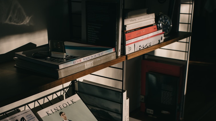 A wooden bookshelf filled with various books and magazines, some stacked and others neatly lined. An old-fashioned iPod and a pair of earbuds rest on top of a pile of books on the upper shelf. There is a clock and some decorative items also present, contributing to a vintage and intellectual atmosphere, highlighted by soft, warm lighting and shadows.