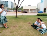 Three students are in a park-like setting, with two of them sitting on the grass and embracing, while a third stands nearby leaning against a tree, reading a book. The scene includes modern buildings in the background and motorcycles parked in the distance.