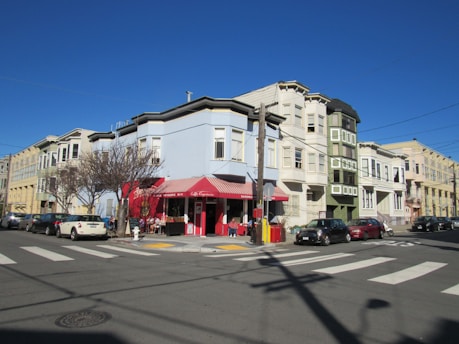 A street corner features a colorful cafe with red accents and a striped awning. People are sitting at tables outside, surrounded by parked cars and a few leafless trees. The surrounding buildings are a mix of light blue, green, and beige, with a clear blue sky above.