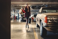A person is walking in a dimly lit parking garage carrying multiple shopping bags. They are dressed stylishly, wearing sunglasses, and walking near a vintage car parked on the right side.