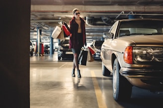 A person is walking in a dimly lit parking garage carrying multiple shopping bags. They are dressed stylishly, wearing sunglasses, and walking near a vintage car parked on the right side.