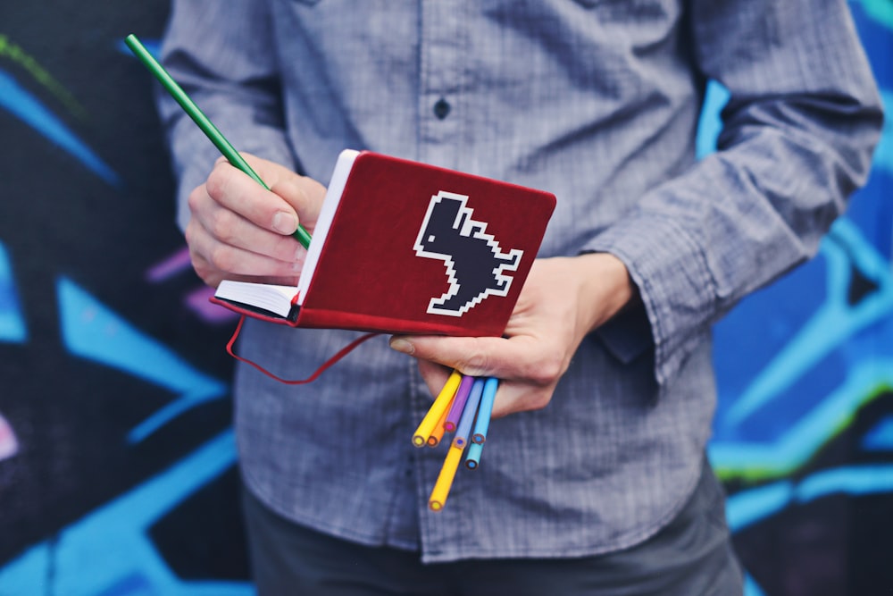 A person taking notes in a red notebook with a dinosaur on its cover while standing