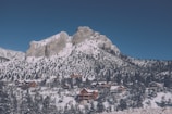 A majestic mountain covered in snow rises above a forested landscape. At the base of the mountain, a cluster of wooden cabins and lodges nestle among snow-laden trees, creating a serene alpine village scene.