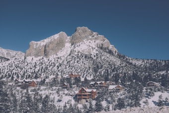 A majestic mountain covered in snow rises above a forested landscape. At the base of the mountain, a cluster of wooden cabins and lodges nestle among snow-laden trees, creating a serene alpine village scene.