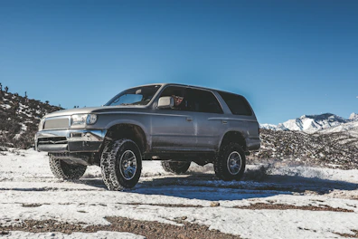 A sturdy silver SUV driving on a rugged mountain trail with a clear blue sky.
