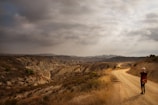 A lone traveler walks along a winding dirt path that stretches through a barren, rugged landscape under a cloudy sky. The ground is dry and rocky, with some scrubby vegetation dotting the path. The scene evokes a sense of isolation and adventure.