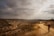 A lone traveler walks along a winding dirt path that stretches through a barren, rugged landscape under a cloudy sky. The ground is dry and rocky, with some scrubby vegetation dotting the path. The scene evokes a sense of isolation and adventure.