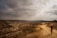 A lone traveler walks along a winding dirt path that stretches through a barren, rugged landscape under a cloudy sky. The ground is dry and rocky, with some scrubby vegetation dotting the path. The scene evokes a sense of isolation and adventure.