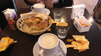 A table holds a white cup of coffee, a wicker basket with croissants and pastries, two glasses of water, and a carton of chocolate milk. A few sugar packets and a napkin holder are also present. The setting appears to be a cafe or breakfast spot.