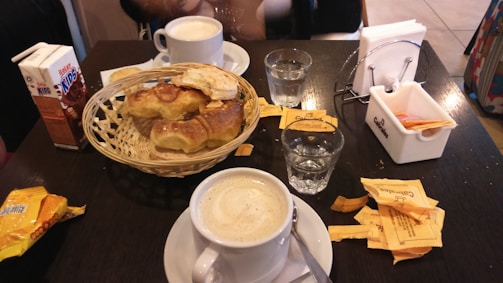 A table holds a white cup of coffee, a wicker basket with croissants and pastries, two glasses of water, and a carton of chocolate milk. A few sugar packets and a napkin holder are also present. The setting appears to be a cafe or breakfast spot.
