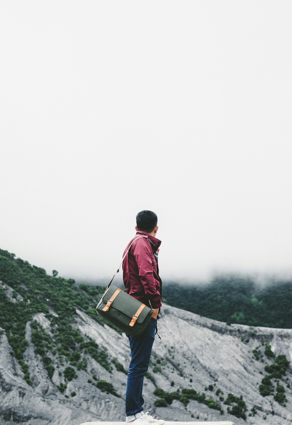 man carrying black crossbody bag while standing on platform overlooking hill under white sky at daytime