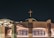 A modern church building with a cross illuminated on top. The structure features large glass doors and arched roofing, with stone and brick elements creating a symmetrical facade. The surrounding area is well-lit against the night sky, highlighting the architectural details of the entrance.