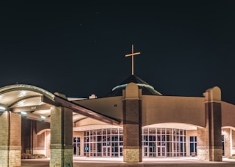 A modern church building with a cross illuminated on top. The structure features large glass doors and arched roofing, with stone and brick elements creating a symmetrical facade. The surrounding area is well-lit against the night sky, highlighting the architectural details of the entrance.