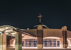 A modern church building with a cross illuminated on top. The structure features large glass doors and arched roofing, with stone and brick elements creating a symmetrical facade. The surrounding area is well-lit against the night sky, highlighting the architectural details of the entrance.