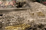 A close-up of a clear plastic storage box containing various earring hooks, clasps, and metal findings. The compartments are filled with metallic items, with a mixture of silver and gold tones, surrounded by a blurred background of more crafting materials.