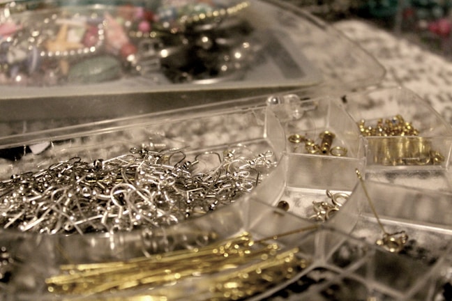 A close-up of a clear plastic storage box containing various earring hooks, clasps, and metal findings. The compartments are filled with metallic items, with a mixture of silver and gold tones, surrounded by a blurred background of more crafting materials.