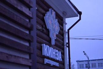 A logo sign attached to the wooden exterior of a building, featuring a stylized design in light and dark colors. Below the logo, text appears in a bold font. The background includes part of the building's facade and a view of street elements such as power lines and a street lamp, under a blue-tinged sky.