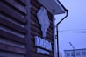 A logo sign attached to the wooden exterior of a building, featuring a stylized design in light and dark colors. Below the logo, text appears in a bold font. The background includes part of the building's facade and a view of street elements such as power lines and a street lamp, under a blue-tinged sky.