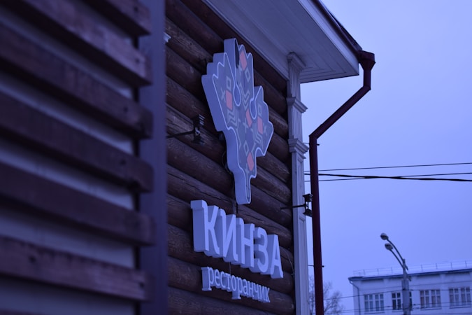A logo sign attached to the wooden exterior of a building, featuring a stylized design in light and dark colors. Below the logo, text appears in a bold font. The background includes part of the building's facade and a view of street elements such as power lines and a street lamp, under a blue-tinged sky.