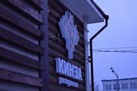 A logo sign attached to the wooden exterior of a building, featuring a stylized design in light and dark colors. Below the logo, text appears in a bold font. The background includes part of the building's facade and a view of street elements such as power lines and a street lamp, under a blue-tinged sky.