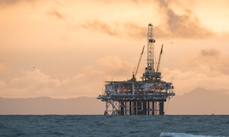 An offshore oil rig stands in the ocean against a backdrop of distant mountains and a dramatic sky, with a warm glow and scattered clouds.